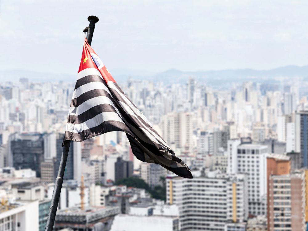 Bandeira do estado de São Paulo tremulando em destaque, com a paisagem urbana da cidade ao fundo.